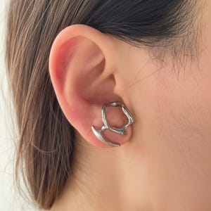 May include: A silver ear cuff with an abstract, geometric design. The cuff wraps around the ear, featuring a curved, pointed element and an open, irregular shape. The jewelry is worn on the ear.