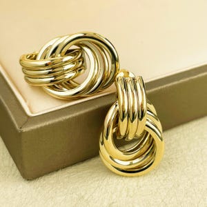May include: Gold-coloured earrings with a knotted design. The earrings feature multiple interlocking loops, creating a bold and textured appearance. The jewellery is displayed on a neutral surface.