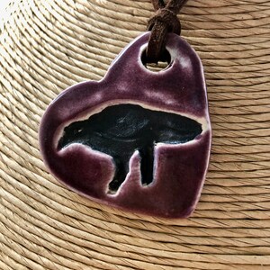 May include: A purple heart-shaped ceramic pendant with a black silhouette of a bear inside. The pendant is on a brown leather cord.