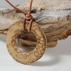 May include: A brown ceramic ring pendant with a textured surface hangs from a brown leather cord. The pendant is on a piece of driftwood.