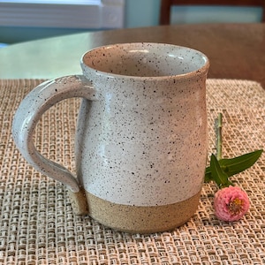May include: A ceramic mug with a speckled white glaze on the upper portion and a textured, unglazed tan base. The mug features a curved handle and a wide rim. A small pink zinnia flower with green leaves is positioned next to the base of the mug.