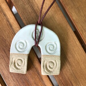 May include: A light blue ceramic pendant shaped like an arch with a brown leather cord. The pendant has a textured surface with a swirl design.