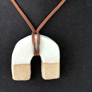 May include: A white ceramic pendant shaped like an arch with a brown suede cord. The pendant has a light brown glaze on the bottom.