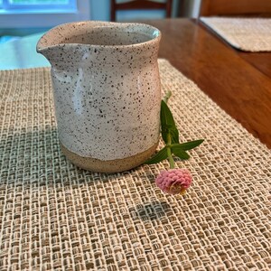May include: A ceramic creamer pitcher with a speckled white glaze on the upper portion and a natural tan clay base. The pitcher features a small pouring spout and a rounded body. A single pink zinnia flower with green leaves rests beside it.