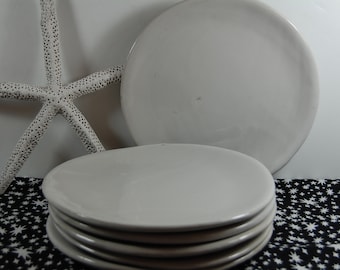 May include: A stack of six white ceramic plates, with a larger white ceramic plate behind them. The plates are on a black and white fabric with a star pattern.