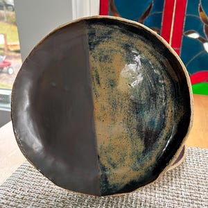 May include: A round ceramic plate featuring a dual-tone finish. The left side is a smooth, matte dark brown, while the right side displays a glossy, speckled glaze in shades of tan, blue, and black with a textured, mottled appearance.