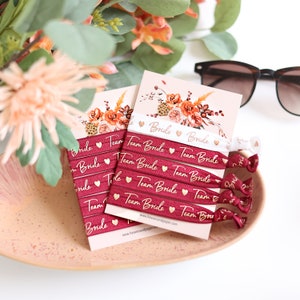 Könnte beinhalten: Ein Set aus burgunderfarbenen elastischen Haargummis mit goldener Schrift, die "Team Bride" und ein kleines goldenes Herz zeigt. Die Haargummis sind auf einer weißen Karte mit einem Blumendesign und der Website-Adresse "www.heavenandpaper.com" verpackt.