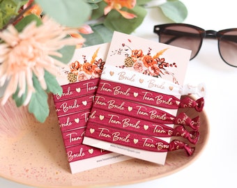 May include: A set of burgundy elastic hair ties with gold foil lettering that says "Team Bride" and a small gold heart. The hair ties are packaged on a white card with a floral design and the website address "www.heavenandpaper.com".