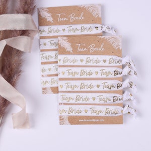 Könnte beinhalten: Ein Set weißer Gummibänder für Haare mit goldener Folienbeschriftung, auf der "Team Bride" steht. Die Haargummis sind auf einer braunen Karte mit dem gleichen Text verpackt.