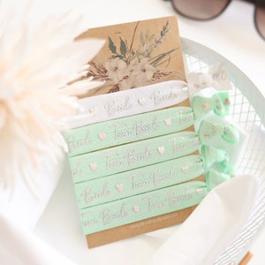 Könnte beinhalten: Ein Set mit sechs mintgrünen und weißen Gummibändern für Haare mit silbernen Glitzerbuchstaben. Die Haargummis sagen "Bride" und "Team Bride".