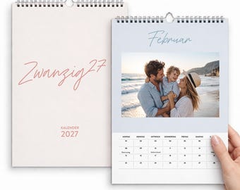 May include: A wall calendar for the year 2027 with a spiral binding. The cover features the text Zwanzig 27 and Kalender 2027. The open page displays the month of Februar with a photograph of three people on a beach and a grid calendar.