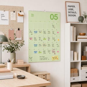 Op de afbeelding: Een lichtgroene wandkalender voor mei 2027 met een rasterindeling, handgeschreven notities, afspraken en kleine kleurrijke stickers. De kalender hangt aan een muur naast een pegboard, een bureaulamp en een witte boekenplank.