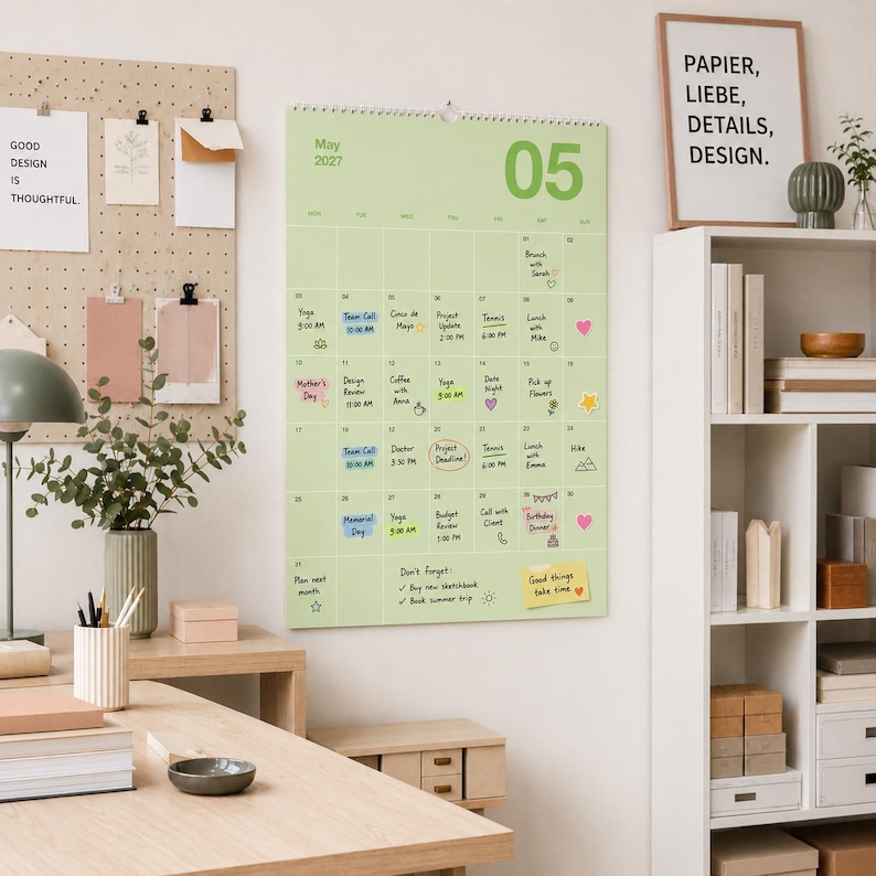 Op de afbeelding: Een lichtgroene wandkalender voor mei 2027 met een rasterindeling, handgeschreven notities, afspraken en kleine kleurrijke stickers. De kalender hangt aan een muur naast een pegboard, een bureaulamp en een witte boekenplank.