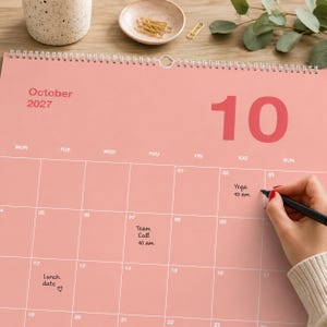 Op de afbeelding: Een wandkalender voor oktober 2027 met een effen roze achtergrond en witte rasterlijnen. De kalender bevat de tekst October 2027 en het cijfer 10 in een groot lettertype. Handgeschreven notities zijn onder andere Yoga 10 am, Team Call 10 am en Lunch date.