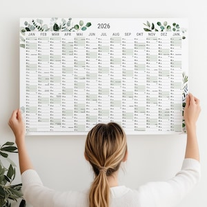 Könnte beinhalten: Ein großer Wandkalender 2026 mit weißem Hintergrund und grünen botanischen Akzenten. Das Kalendarium ist hellgrün. Die Monate sind oben aufgeführt, mit dem Jahr 2026 prominent angezeigt.