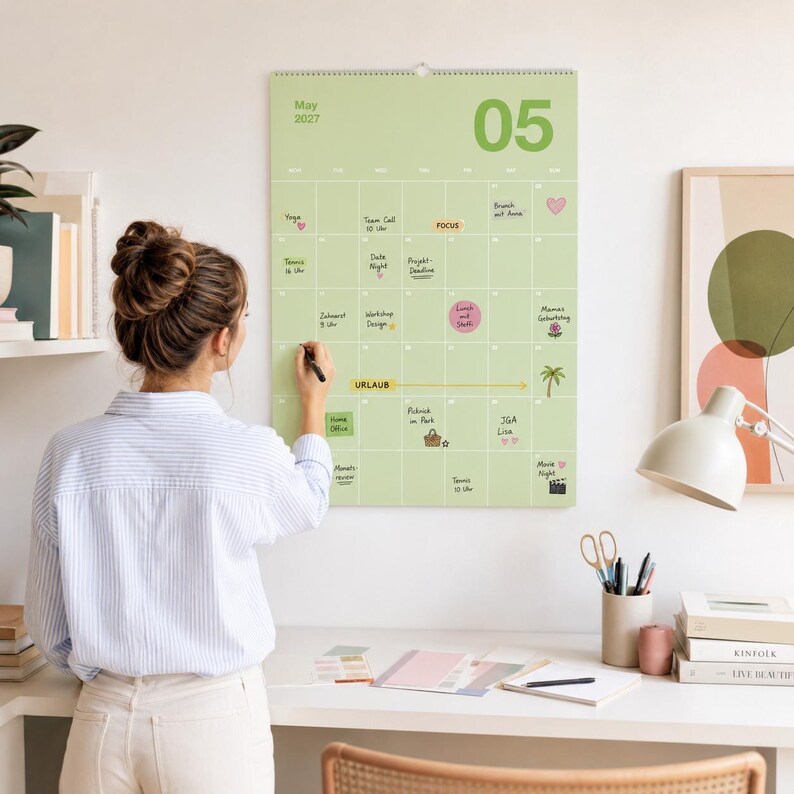 Op de afbeelding: Een lichtgroene wandkalender voor mei 2027 met een rasterindeling, handgeschreven notities, kleine iconen en de tekst "URLAUB" die over meerdere dagen loopt. De kalender hangt aan een muur boven een wit bureau met kantoorbenodigdheden en boeken.