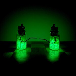 Può includere: Due piccole bottiglie di vetro con tappi neri e contenuto verde brillante sono collegate da un sottile filo metallico. Le bottiglie poggiano su una superficie a scacchi, illuminate dalla luce verde, contro uno sfondo verde scuro.