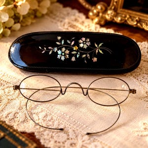 May include: A pair of oval-shaped metal eyeglasses with thin frames and a matching black oval case featuring an inlaid iridescent floral mother-of-pearl design on the lid. The glasses have a high bridge and thin wire temples.