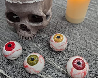 Può includere: Una scultura di teschio grigio cavo e usurato posizionata accanto a una candela a colonna LED gialla luminosa. Cinque oggetti di scena sferici a forma di bulbo oculare sono disposti davanti, con sclera bianca con venature rosse e iridi rosse, oro, oliva e grigie.