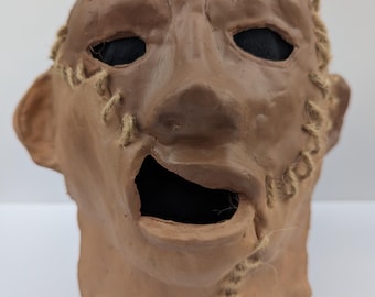 Può includere: Una maschera scolpita color marrone di una testa umana con una bocca larga e aperta e orbite nere profonde. La superficie è strutturata con cuciture visibili unite da uno spago spesso color beige che attraversa la fronte e il mento.
