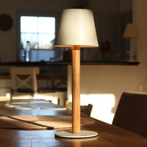 May include: A contemporary table lamp featuring a cream lampshade and a wooden stem. The lamp is set on a white circular base. The lamp is on a wooden table in a well-lit room.