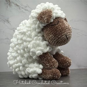 May include: A handmade, plush sheep toy with a white, fluffy body and brown head, ears, and legs. The sheep is sitting down, with its head tilted slightly downward. The text "Hatched Crocheted Creations" is at the bottom.