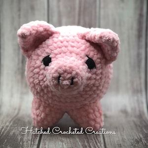 May include: A pink crocheted pig with black eyes. The pig is standing on a wooden surface.