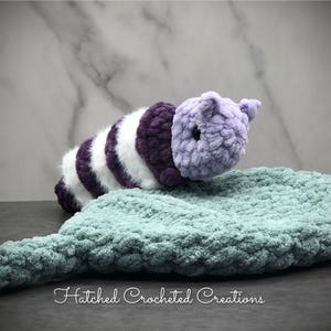 May include: A handmade crochet caterpillar toy in shades of purple, white, and teal. The caterpillar is resting on a teal-colored leaf-shaped crochet mat. The text "Hatched Crocheted Creations" is at the bottom.