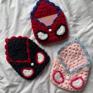 Puede incluir: Tres estuches de ganchillo hechos a mano con forma de sobre, cada uno con un pequeño botón blanco y dos detalles en forma de ojos blancos. Un estuche es rojo con borde azul, otro negro con borde rojo y otro rosa claro con borde azul claro.