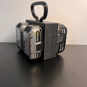 May include: A black 3D-printed battery holder for Ryobi 18V batteries. The holder has a handle for easy carrying and is designed to hold multiple batteries. The Ryobi logo is visible on the side of the holder. The batteries are black and gray.