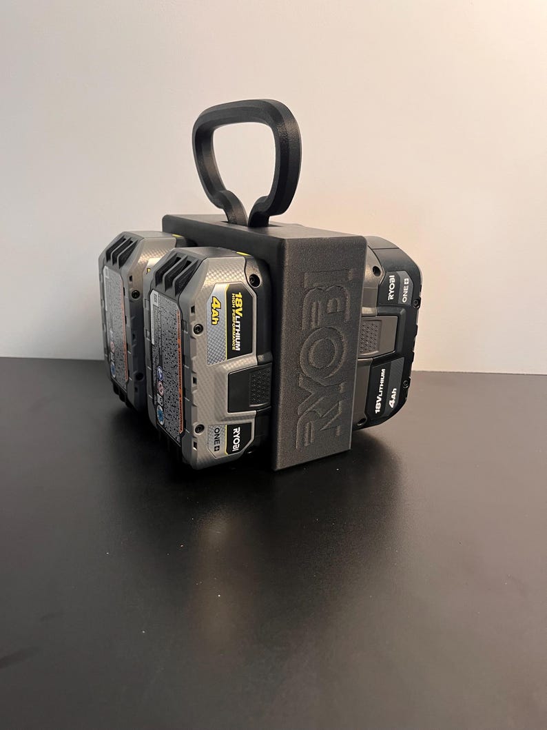 May include: A black 3D-printed battery holder for Ryobi 18V batteries. The holder has a handle for easy carrying and is designed to hold multiple batteries. The Ryobi logo is visible on the side of the holder. The batteries are black and gray.