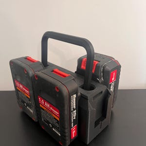 May include: A black 3D-printed battery holder with a handle, holding three black and red Bauer 20V Hypermax Lithium-Ion 1.5 AH batteries. The batteries have red accents and white text. The holder is on a black surface.