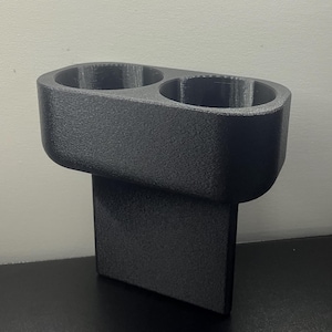 May include: A black, 3D-printed car cup holder with two circular openings. The cup holder has a textured surface and a rectangular base for secure placement. The design is simple and functional.