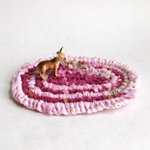 May include: A small, oval-shaped rug in shades of pink and burgundy, with a miniature deer figurine standing on top. The rug appears to be handmade, with a braided or woven texture. The deer is brown with white accents.