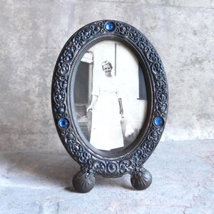 May include: An oval, antique-style picture frame with a black and white photograph. The frame is dark gray with ornate scrollwork and three blue, round embellishments. The frame has a stand with two decorative feet.