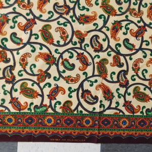 Op de afbeelding: Een crèmekleurige stof met een paisley-patroon in tinten groen, rood en bruin. De stof heeft een rand met een herhalend patroon van rood, groen en goud. De stof heeft de tekst "VERITABLE JAVA HOLLANDAIS 41618" gedrukt op de onderkant.