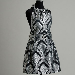 May include: A black and silver damask apron with a halter neck and a belt. The apron has a ruffled skirt.