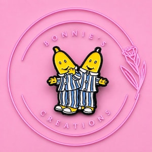 May include: A metal enamel pin featuring two yellow banana characters wearing blue and white striped pyjamas. The pin is centred within a circular pink frame with the text BONNIE'S CREATIONS and a small rose illustration on the right side.