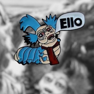 May include: A blue and red enamel pin featuring a cartoon character with a speech bubble that says "Ello". The character has blue hair, a red scarf, and a blue body with a segmented design.
