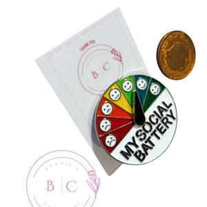 May include: A white enamel pin with a rainbow gauge that reads "My Social Battery". The gauge has a black needle and is surrounded by a circle of six different colored smiley faces, ranging from happy to sad.