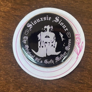 May include: A circular pin-back button featuring a black and white graphic of Siouxsie Sioux. The text reads Siouxsie Sioux and The Original 80's Goth Mother Queen, with small coffin icons on the sides. The button is mounted on a white circular card.
