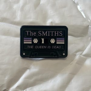 Puede incluir: Un pin de esmalte verde oscuro con forma de cinta de cassette. El pin presenta el nombre de la banda "The Smiths" y el título del álbum "The Queen Is Dead" en letras blancas. El diseño incluye rayas moradas y blancas.