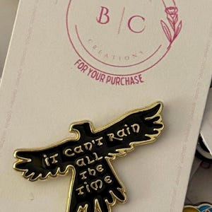 Puede incluir: Pin de esmalte negro y dorado con un pájaro negro estilizado con el texto "it can't rain all the time".