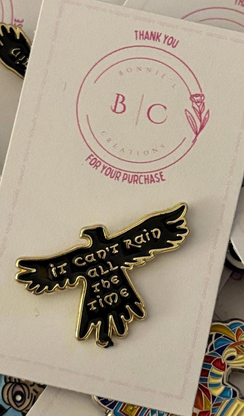 May include: Gold and black enamel pin featuring a stylised black bird with the text "it can't rain all the time".