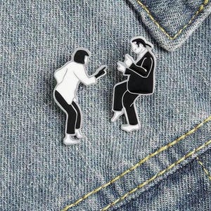 以下が含まれることがあります： 男性と女性が踊っている様子を描いた白黒のエナメルピン2つ。男性はスーツを着ており、女性はドレスを着ています。ピンは青いデニムの背景に置かれています。