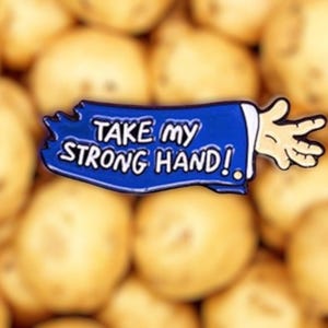 Puede incluir: Un pin de esmalte azul con una mano de dibujos animados extendida y el texto "TAKE MY STRONG HAND!" en una pancarta azul. El pin está enfocado contra un fondo borroso de patatas.