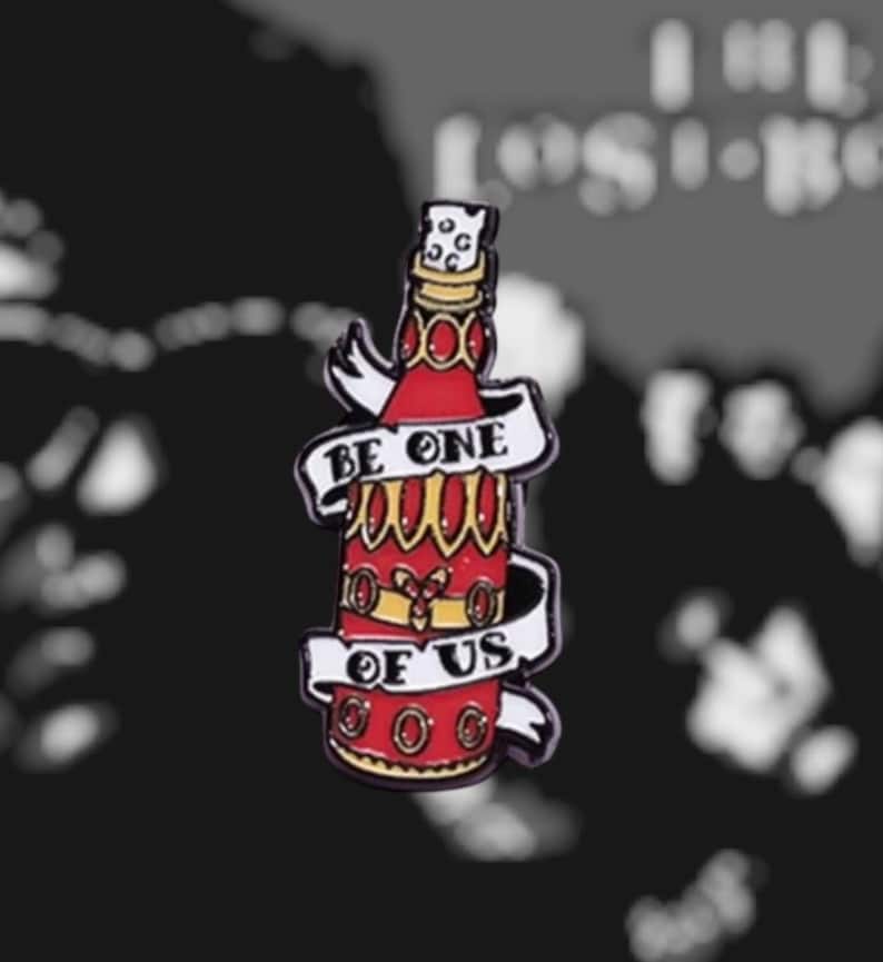 Puede incluir: Un pin de esmalte en forma de botella roja y dorada con las palabras "BE ONE OF US" en una pancarta blanca. El pin tiene un dise&ntilde;o decorativo con detalles en rojo y dorado, y una parte superior similar a un corcho.