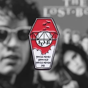 May include: Red and white enamel pin shaped like a coffin with a bridge and people on it. The pin says "You'll never grow old, You'll never die."