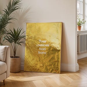 Peut inclure: Intérieur de salon lumineux avec un canapé beige, des coussins et une couverture. Une œuvre d'art encadrée dorée avec le texte "Your custom text here" est appuyée contre le mur. Des plantes en pot et une fenêtre complètent la scène.