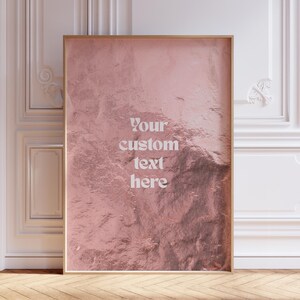 Peut inclure: Une affiche encadrée avec une surface texturée rose doré. L'affiche porte le texte "Your custom text here" en blanc. Le cadre est de couleur marron clair. Le fond présente des boiseries blanches et un parquet.